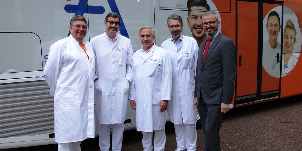Vier Ärzte in Kitteln und ein Mann im Anzug stehen vor einem großen Bus, welcher Werbung für die Augustinus Gruppe trägt und gucken lächelnd in die Kamera.