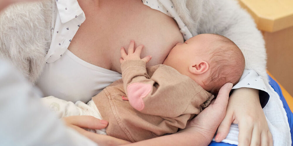 Eine junge Mutter sitzt und stillt ihr Neugeborenes hierbei steht eine Frau neben ihr und schaut auf sie runter.