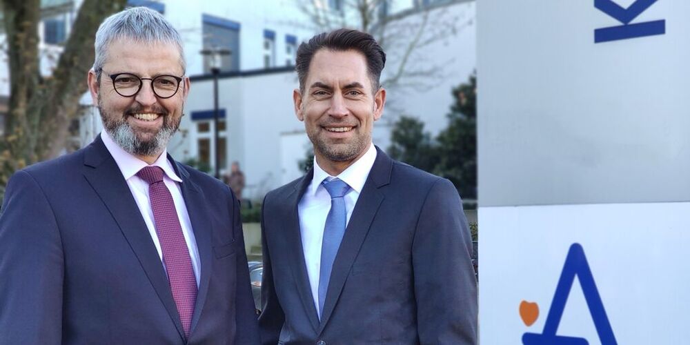 Zwei Männer in Anzügen stehen vor einem Eingangsschild mit dem Augustinus Logo. 