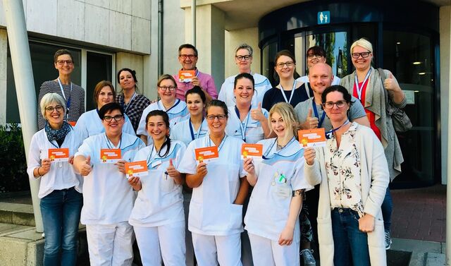 Mitarbeiterinnen und Mitarbeiter der Pflege stehen für ein Gruppenbild zusammen und halten eine Werbekarte in der Hand, welche sie in die Kamera zeigen. Alle schauen lächelnd in die Kamera.